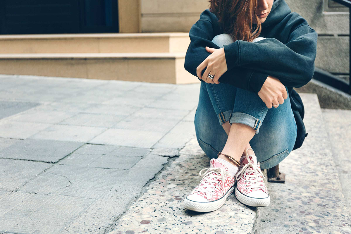 A young teen sits with they arms around their knees and looks off into the distance